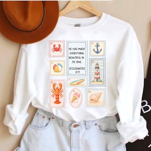 May include: A white long-sleeve sweatshirt featuring a graphic print of various postage stamps. The stamps display a crab, a beach ball, a lobster, an ice cream cone, an anchor, a lighthouse, a surfboard, and a seashell. Text reads: HE HAS MADE EVERYTHING BEAUTIFUL IN ITS TIME. ECCLESIASTES 3:11.