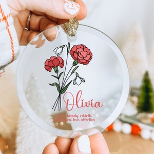 Puede incluir: Adorno navide&ntilde;o circular de cristal transparente con un dise&ntilde;o floral de claveles rojos y campanillas blancas. El nombre "Olivia" est&aacute; escrito en cursiva, con las palabras "esperanza, belleza, renacimiento, admiraci&oacute;n, amor, afecto."