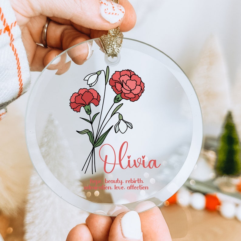 Puede incluir: Adorno navide&ntilde;o circular de cristal transparente con un dise&ntilde;o floral de claveles rojos y campanillas blancas. El nombre "Olivia" est&aacute; escrito en cursiva, con las palabras "esperanza, belleza, renacimiento, admiraci&oacute;n, amor, afecto."