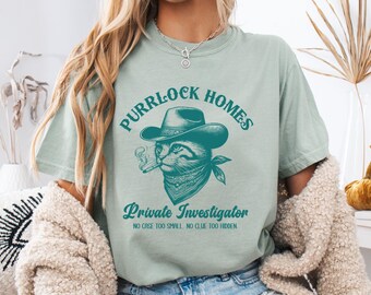 May include: A light green t-shirt with a teal graphic of a cat wearing a cowboy hat and bandana, smoking a cigarette. The text reads "Purrlock Homes, Private Investigator, No Case Too Small, No Clue Too Hidden."