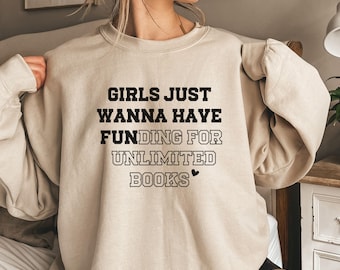 May include: A beige crewneck sweatshirt featuring black text that reads GIRLS JUST WANNA HAVE FUNDING FOR UNLIMITED BOOKS with a small black heart icon. The garment has long sleeves and a ribbed collar, cuffs, and hem, providing a casual look.