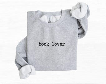 Puede incluir: Una sudadera gris claro de cuello redondo con el texto "book lover" en fuente negra estilo máquina de escribir en el pecho. La prenda tiene mangas largas con puños acanalados y un forro interior de felpa blanca visible en el cuello y puños.