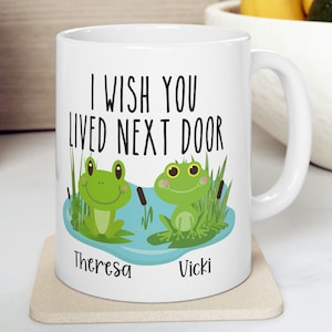 May include: White ceramic mug with a green frog illustration and the text "I wish you lived next door". The mug has two names, "Theresa" and "Vicki", written below the frogs.