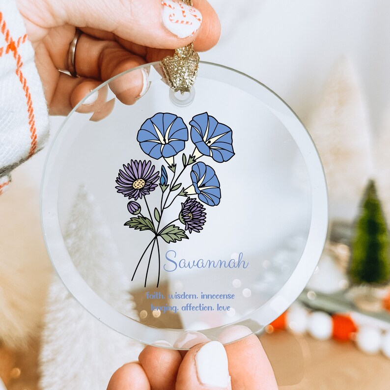 Puede incluir: Un adorno de cristal transparente con un dise&ntilde;o floral con flores azules y moradas. El nombre "Savannah" est&aacute; escrito en una fuente cursiva debajo de las flores, junto con las palabras "fe, sabidur&iacute;a, inocencia, amor, afecto, amor."