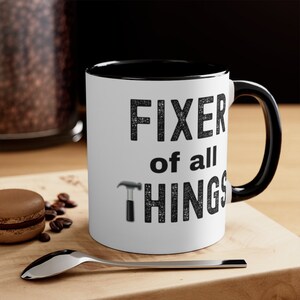 May include: White coffee mug with a black rim and handle. The mug has black text that reads "FIXER of all THINGS" with a small black hammer graphic.