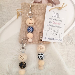 Könnte beinhalten: Zwei Schlüsselanhänger aus Holzperlen mit verschiedenen Anhängern. Ein Schlüsselanhänger hat einen Herzanhänger und der andere einen Ankeranhänger. Beide Schlüsselanhänger haben eine Perle mit einem schwarz-weißen Tiermuster und eine Holzperle mit einem hellblau-weißen Muster. Die Schlüsselanhänger befinden sich auf einem weißen und braunen Stoffhintergrund. Der Text auf der Karte lautet "SORRY, aber wir müssen für immer befreundet bleiben. Du weißt zu viel!"