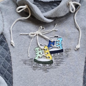 May include: A gray hooded sweatshirt with a decorative patch of two colorful sneakers. The sneakers are blue, yellow, and green with white laces. The sweatshirt has white drawstrings and a quilted patch on the side.