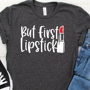 May include: A dark gray t-shirt with the text "But first lipstick" in white lettering and a red lipstick graphic.