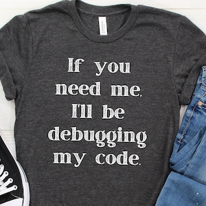 May include: A dark grey t-shirt with the text "If you need me, I'll be debugging my code." printed in white.