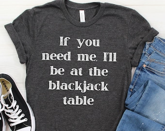 May include: A dark gray t-shirt with the text "If you need me, I'll be at the blackjack table" printed in white.