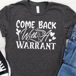 Peut inclure: Un t-shirt gris fonc&eacute; avec le texte "Come Back With A Warrant" en lettres blanches. Le texte est stylis&eacute; avec un c&oelig;ur et un graphique de plante.
