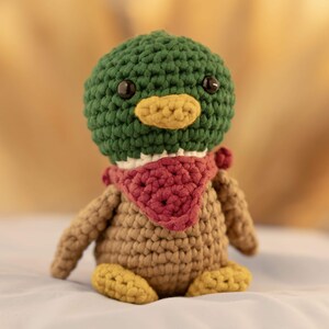 May include: A crocheted duck plush toy with a green head, brown body, and yellow feet. The duck is wearing a red bandana.