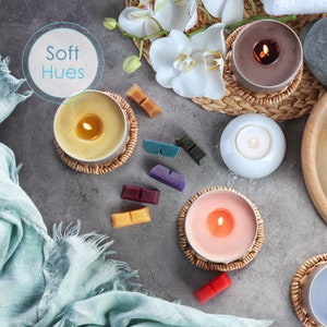 May include: A collection of candles in various colors and sizes, including a yellow candle, a pink candle, a brown candle, and a white candle. The candles are arranged on a gray surface with a blue fabric in the background. The candles are surrounded by small, colorful wax melts.