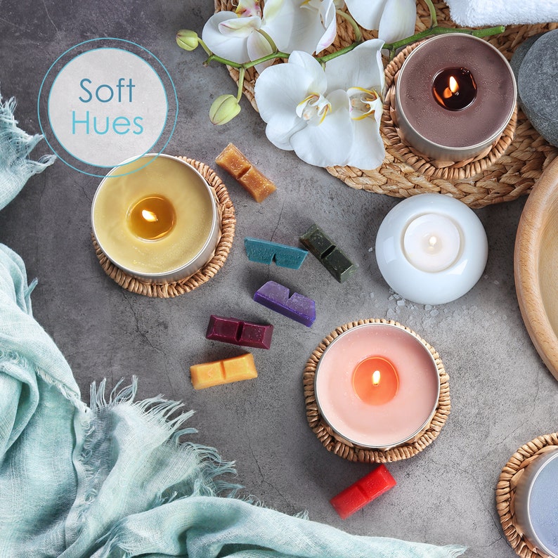 May include: A collection of candles in various colors and sizes, including a yellow candle, a pink candle, a brown candle, and a white candle. The candles are arranged on a gray surface with a blue fabric in the background. The candles are surrounded by small, colorful wax melts.
