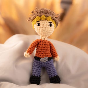 May include: A crocheted doll wearing a brown hat, orange shirt, blue jeans, and black shoes.