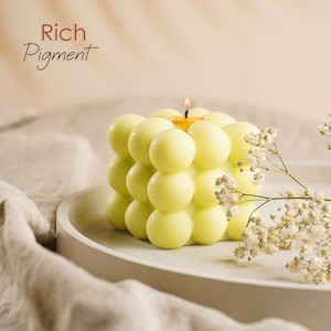 May include: A yellow candle with a textured, bubble-like surface. The candle is lit and sitting on a white plate. The text "Rich Pigment" is visible in the top left corner of the image.