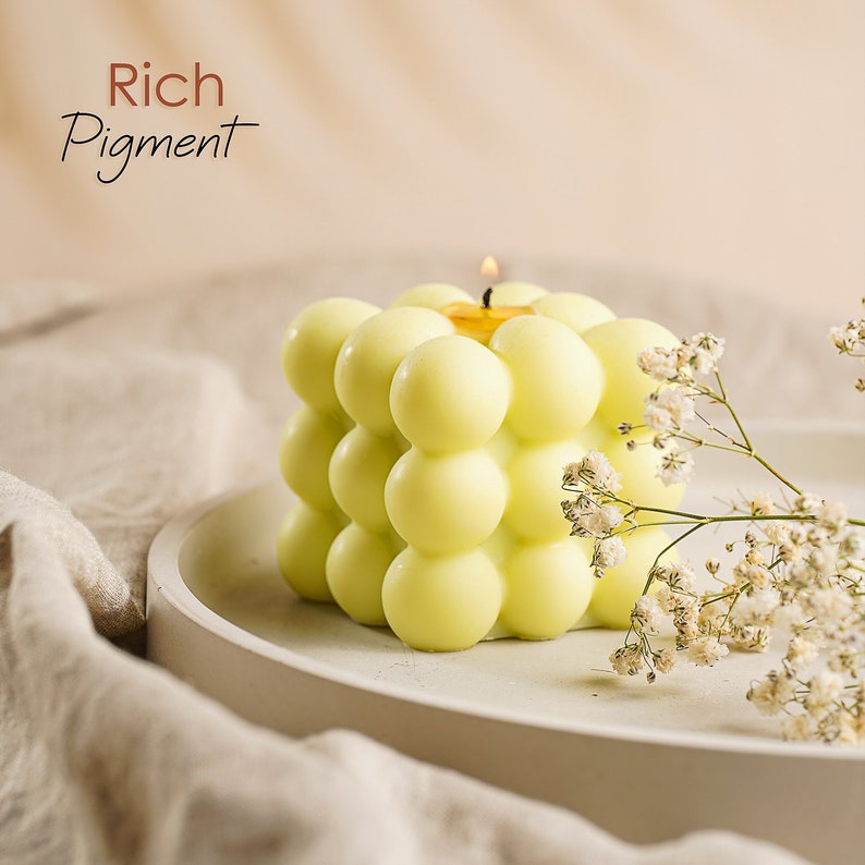 May include: A yellow candle with a textured, bubble-like surface. The candle is lit and sitting on a white plate. The text "Rich Pigment" is visible in the top left corner of the image.