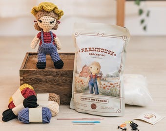May include: A crochet doll wearing overalls and a straw hat stands on a wooden box next to a "Farmhouse Crochet Kit" for beginners. The kit includes yarn in various colors, crochet hooks, and other supplies. The kit features "Dottie the Farmer".