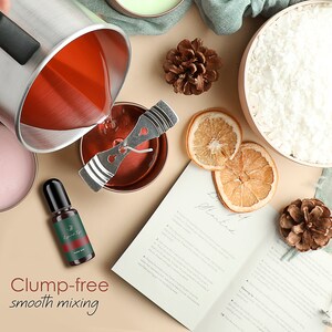 May include: A close-up of a red candle wax being poured into a glass jar with a wick and a metal holder. The wax is being poured from a silver pitcher. There is a bottle of essential oil, a book with instructions, and other decorative items in the background.