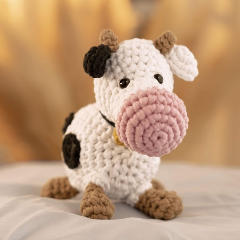 May include: A crocheted stuffed cow toy with white fur, black spots, brown hooves, and a pink nose. The cow has a black and white ear and a bell around its neck.