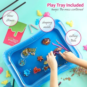 May include: A blue inflatable play tray filled with colorful slime, shaping molds, and cutting tools. The set includes blow straws and a booklet that reads "It's Time For A Slimetastic Adventure!" The text "Play Tray Included keeps the mess contained" is also visible.