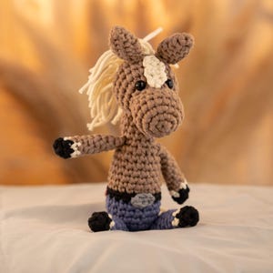 May include: A crocheted brown horse toy with a white mane and a white stripe on its face. The horse is wearing blue jeans with a brown belt.