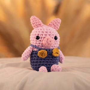 May include: A pink crocheted pig wearing blue overalls with yellow buttons. The pig has black eyes and a pink snout.