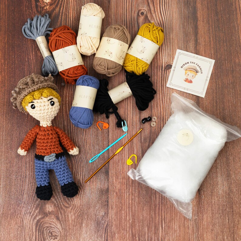 May include: Crochet kit featuring a stuffed toy farmer with a brown hat, orange shirt, and blue jeans. The kit includes yarn in various colors, stuffing, and needles. The kit also includes a card with the text "Dixon the Farmer".