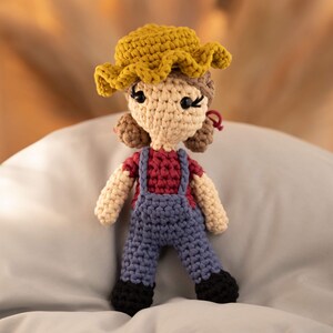 May include: A crocheted doll wearing a yellow hat, a red shirt, and blue overalls. The doll has brown hair and black eyes.