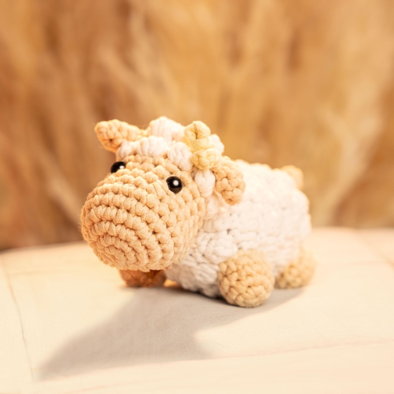 May include: A crocheted white and beige lamb toy with a yellow flower on its head. The toy is lying on a white surface.