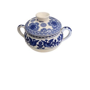 May include: A white and blue ceramic sugar bowl with a lid. The bowl features a blue floral and bird design. The bowl has two handles and a small knob on the lid. The sugar bowl is a decorative kitchen item.