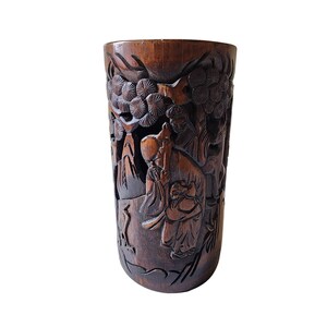 May include: A dark brown, cylindrical wooden vase with intricate carvings depicting a landscape scene with figures and trees. The detailed relief work showcases a traditional Asian art style, with a smooth, polished finish.