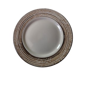 May include: A round, off-white ceramic plate with a concentric, textured rim in shades of brown and cream. The plate has a glossy, smooth center. The design suggests a rustic or natural aesthetic, suitable for serving food.