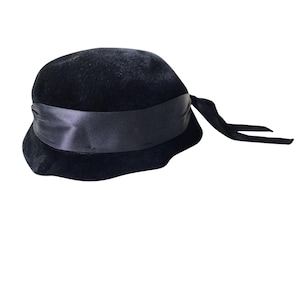 May include: A black cloche hat with a wide satin ribbon band and long, flowing ties. The hat has a rounded crown and a slightly flared brim. The hat is made of a soft, velvety material.