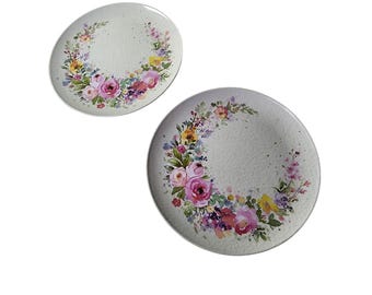 May include: Two white ceramic plates with a floral design. The plates feature a wreath of pink, purple, and yellow flowers with green leaves around the edge.