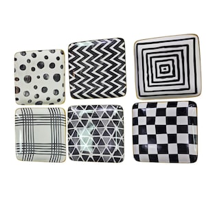 May include: A set of six square ceramic plates featuring distinct black and white patterns including polka dots, zigzags, concentric squares, grid lines, triangles, and a checkerboard design, each finished with a thin gold-toned rim.