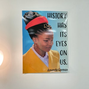 May include: A poster with a quote by Amanda Gorman, "History has its eyes on us." The poster features a portrait of a woman with a red headband and yellow jacket against a blue background.
