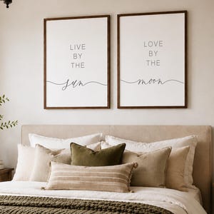 May include: A bedroom featuring a bed with white bedding, a dark green knit throw, and various pillows. Two framed wall prints display the text Live by the sun and Love by the moon. A wooden bench sits at the foot of the bed with a woven basket.