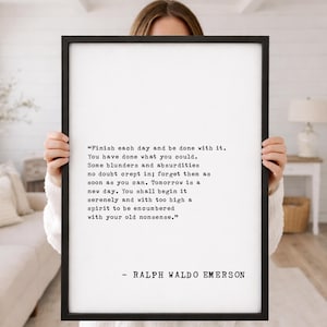 May include: A framed black and white print featuring a quote by Ralph Waldo Emerson. The quote is in a typewriter-style font on a white background. The frame is black and the print is held by a person.
