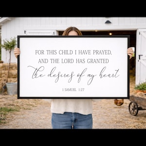 Può includere: Un cartello rettangolare con cornice nera con il testo "FOR THIS CHILD I HAVE PRAYED, AND THE LORD HAS GRANTED the desires of my heart 1 SAMUEL 1:27" in scrittura nera. Il cartello è tenuto da una persona davanti a un fienile bianco.