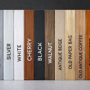 May include: A collection of twelve rectangular wooden planks arranged vertically, each featuring a distinct finish. The labels printed on the planks are Dark Grey, Light Grey, Silver, White, Cherry, Black, Walnut, Antique Beige, Old Paper Bag, Old Antique Coffee, Cashmere, and Natural.