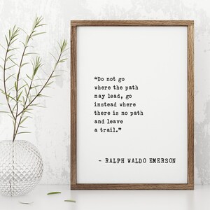 May include: A framed print with a quote by Ralph Waldo Emerson: "Do not go where the path may lead, go instead where there is no path and leave a trail." The print is in a brown wooden frame. A clear glass vase with greenery is to the left.