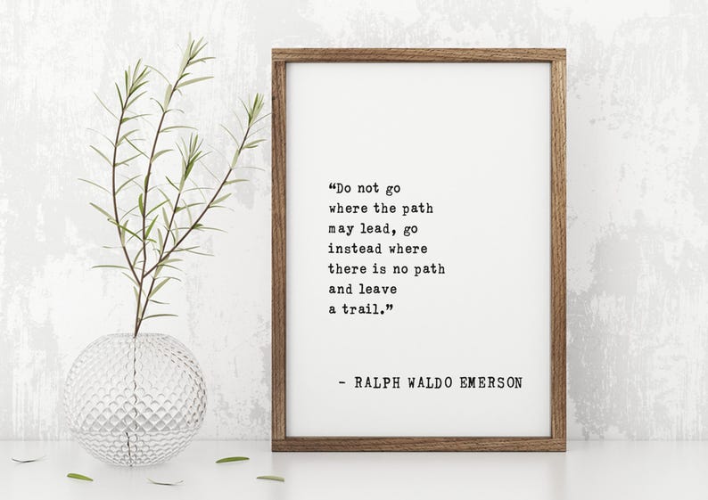 May include: A framed print with a quote by Ralph Waldo Emerson: "Do not go where the path may lead, go instead where there is no path and leave a trail." The print is in a brown wooden frame. A clear glass vase with greenery is to the left.