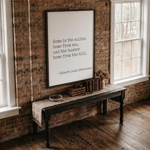 May include: A rectangular wall sign with a dark wood frame featuring the black printed text: Home is the sailor, home from sea, and the hunter home from the hill. - Robert Louis Stevenson. The sign is displayed on a brick wall above a dark wooden table.