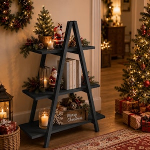 May include: A dark gray wooden A-frame shelf with three tiers, decorated with Christmas items including a small lit tree, Santa figurines, candles in glass holders, books, and a wooden crate with the text Merry Christmas printed on the front.