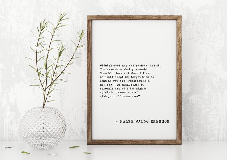 May include: A framed print with a quote by Ralph Waldo Emerson. The text is in black on a white background, set within a wooden frame. A clear glass vase with a plant is to the left of the frame.
