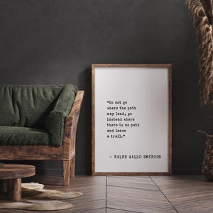 May include: A framed print with a quote by Ralph Waldo Emerson: "Do not go where the path may lead, go instead where there is no path and leave a trail." The print is in a wooden frame, leaning against a dark wall.