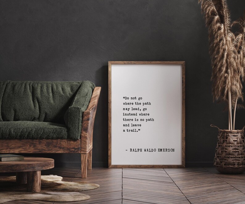 May include: A framed print with a quote by Ralph Waldo Emerson: "Do not go where the path may lead, go instead where there is no path and leave a trail." The print is in a wooden frame, leaning against a dark wall.