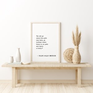 May include: A framed print with the quote "Do not go where the path may lead, go instead where there is no path and leave a trail." by Ralph Waldo Emerson. The print is displayed on a light wood bench with decorative vases and dried plants.