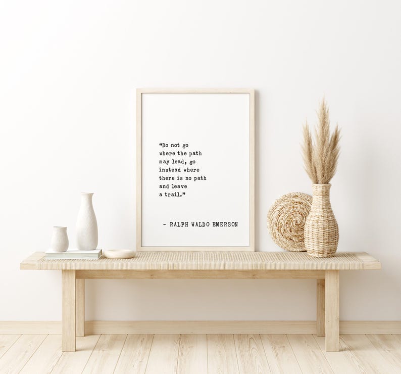 May include: A framed print with the quote "Do not go where the path may lead, go instead where there is no path and leave a trail." by Ralph Waldo Emerson. The print is displayed on a light wood bench with decorative vases and dried plants.